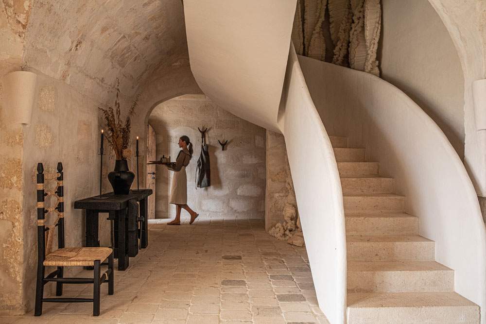 A vaulted stone hallway with a curved plaster staircase, a dark timber table and rush chair against bare limestone walls.