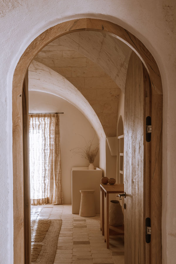 An arched timber doorway looking into a vaulted room, linen curtain drawn across a window and pale stone floor tiles catching the light.