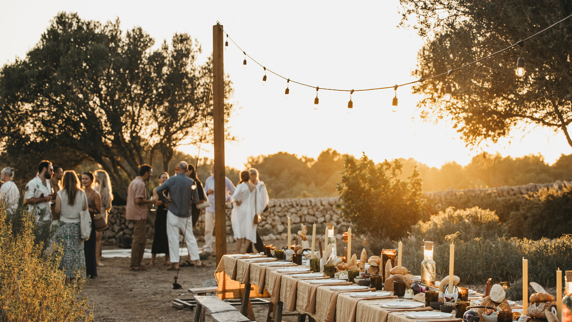 A long communal table dressed with linen and candles beside a dry-stone wall at sunset, guests standing together under a string of lights.
