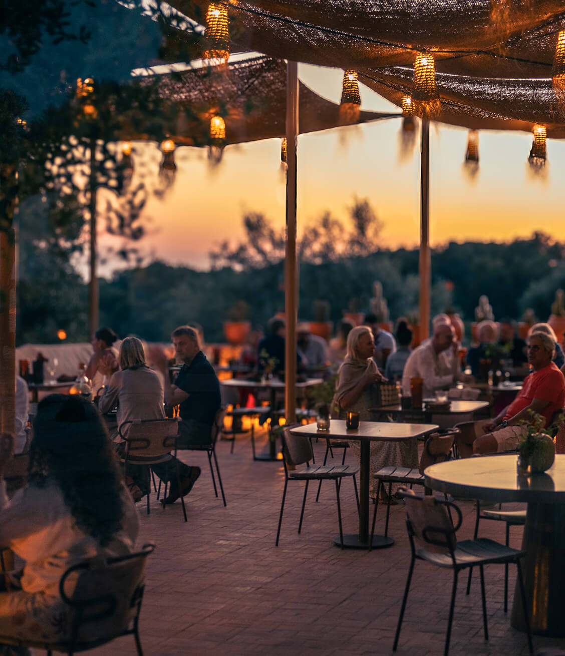 Guests eating on the terrace at dusk under woven shades and hanging lanterns, the sky orange behind the trees.