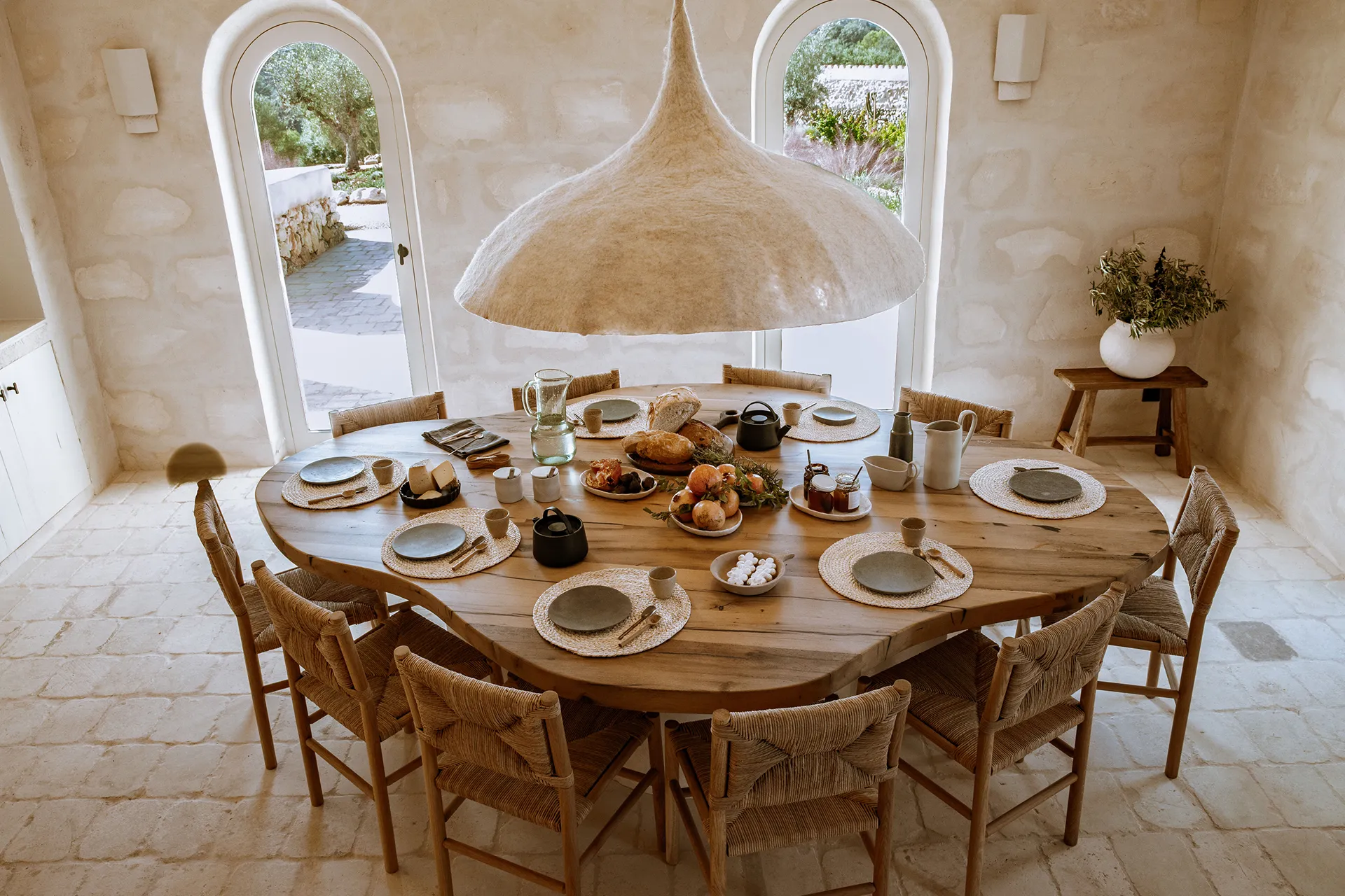 A round timber table laid for eight under a large raffia pendant, breakfast set out in the middle and two arched doorways open to the garden.